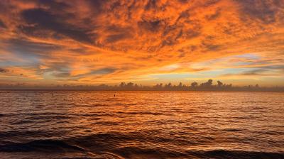 Sunset over the ocean with vibrant orange and yellow hues in the sky, reflecting on the water's surface. Waves gently lap against the shore under dramatic cloud formations.