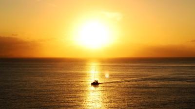 A small boat sails on calm water at sunset, with the sun low on the horizon and golden light reflecting on the sea.